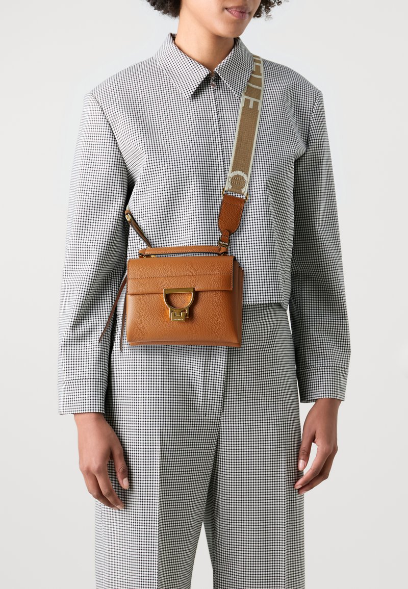 Brun lærcrossbody-veske med strukturert form, gullfarget hardware og justerbar stropp med logo. Mønstret rutete bluse på.