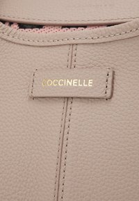 Gros plan sur un cuir beige texturé avec des coutures verticales et une petite étiquette en cuir embossée avec "COCCINELLE" en lettres dorées.