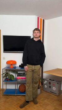 Άνδρας με μαύρο φούτερ με κουκούλα και καφέ παντελόνι cargo στέκεται σε δωμάτιο με τηλεόραση, πορτοκαλί λάμπα, μπάλα μπάσκετ, φυτό και μεταλλική βαλίτσα με την επιγραφή "Tropicana".