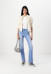 Cardigan lavorato a maglia beige sopra una maglietta bianca, abbinato a jeans a zampa d'elefante blu chiaro. Gli accessori includono una borsa argento e scarpe argentate lucide.