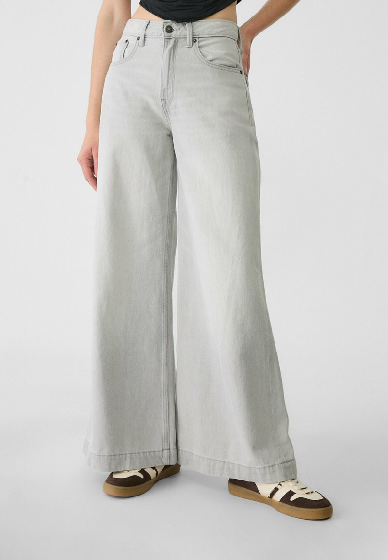 Pantalons en denim gris à jambes larges avec taille haute, dotés de poches avant, d'un zip et de coutures contrastées. Portés avec des baskets décontractées.