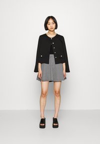 Fekete rövidített dzseki elől gombokkal, párosítva fekete-fehér kutyafog mintás szoknyával. A modellen fekete platform papucsok vannak.