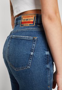 Denim jeans in medium blue with a faded effect. Features a brown brand label on the waistband and two back pockets.
