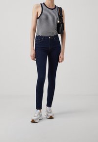 Jeans ajustados de color azul marino con una cintura alta, combinados con una camiseta sin mangas de rayas en blanco y negro. Zapatillas blancas con acentos en gris y beige.