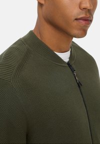 Olive green sweter dzianinowy z zamkiem z przodu, posiadający teksturowany materiał, ribbowe wykończenie kołnierza oraz bezszwowe detale na ramionach.