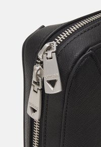 Sort tekstilpose med en glat overflade, der har en sølvmetal lynlås og mærkede lynlåsanhængere. Vinklet udsigt fremhæver det elegante design.