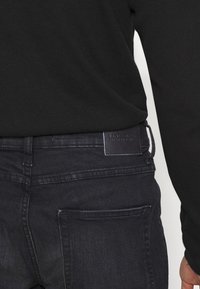 Camisa negra de manga larga combinada con jeans de mezclilla oscuros, que presentan un parche de cuero en la cintura. Los jeans muestran una textura suave y una costura sutil.