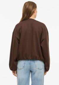 Pruun ülisuur pusa soonikkoes äärega, pikkade varrukatega ja pehme tekstuuriga, mis on kombineeritud hele siniste kõrge vöökohaga teksapükstega.