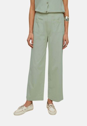 Mujer con pantalones anchos de pierna en verde claro con bolsillos parche delanteros, combinados con zapatos tipo mocasín beige y una pulsera de brazalete plateada.