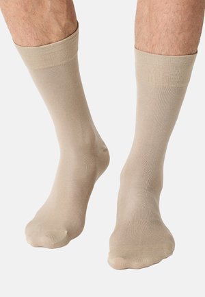 Beine, die beige Knöchelsocken mit glatter Textur tragen und auf einem weißen Hintergrund stehen.