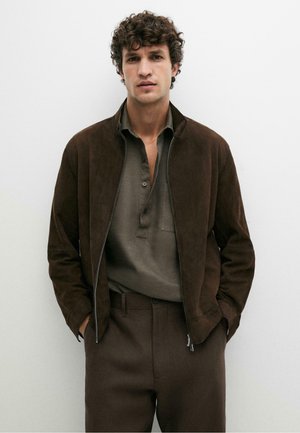 Man met krullend haar, die een donkerbruine suède jas draagt, een grijs overhemd met kraag en een bruine broek, staand met handen in de zakken.
