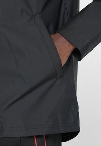 Czarna wodoodporna kurtka o gładkiej fakturze, posiadająca boczną kieszeń i dopasowane mankiety. Szczegóły szwów widoczne wzdłuż dolnego brzegu.