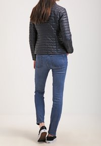 Kobieta w czarnej pikowanej kurtce, niebieskich obcisłych dżinsach i czarno-białych sneakersach, stojąca tyłem do aparatu.