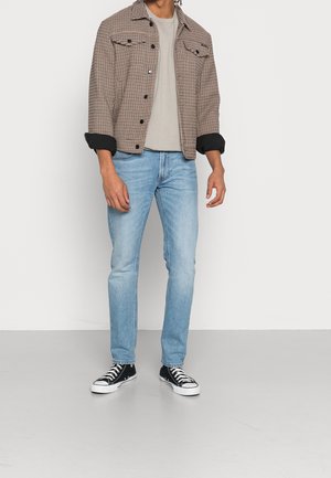 Mand iført en beige ternet jakke over en grå t-shirt, lyseblå jeans og sorte høje sneakers, stående mod en ensfarvet baggrund.