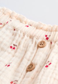 Tissu crème clair avec une surface texturée et froissée, orné d'un imprimé cerises rouges et de boutons en bois brun. Détail de taille à volants.