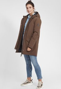 Brun isolert parka-jakke med hette, sidelommer og trykknapper, kombinert med en lyseblå skjorte og denimjeans.