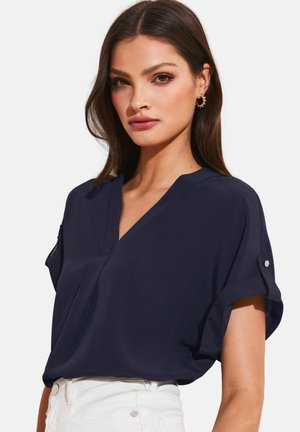 Frau mit glatten braunen Haaren, die goldene Creolen, eine marineblaue V-Ausschnitt-Bluse mit hochgekrempelten Ärmeln und weiße Hosen vor weißem Hintergrund trägt.