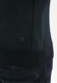 Donker gebreide trui met ribbels aan de onderkant en een subtiel chevronpatroon, gedragen over donkere spijkerbroek, waarbij de textuur van de stof en een klein logo-detail worden benadrukt.
