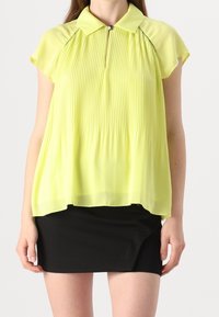 Blusa plissettata verde lime con colletto, maniche corte e dettaglio con zip frontale, abbinata a una gonna nera. Tessuto leggero e fluente.