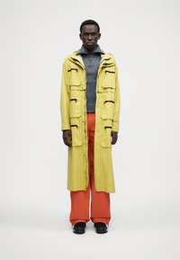 Casaco amarelo longo com múltiplos bolsos, combinado com calças largas laranjas. O casaco apresenta pormenores em zíper preto e um capuz.