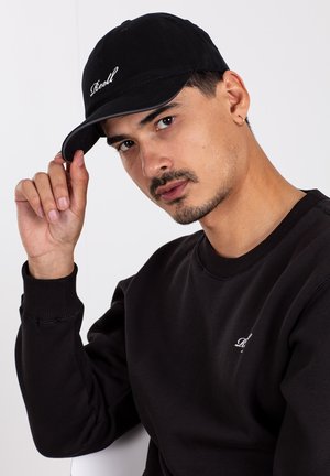 Schwarze Baumwollmütze mit gebogenem Schild und gesticktem weißen Text. Getragen von einer Person in einem schwarzen Sweatshirt, die vor einem hellen Hintergrund sitzt.