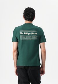 Žalia trumpa rankovė T-maikė su apvalia apykakle, kuriuose ant nugaros puikuojasi baltas tekstas "The Hilfiger Herald" laikraščio stiliumi.