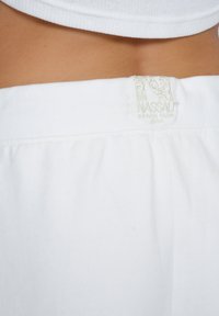 White cotton shorts with an elastic waistband featuring a subtle "NASSAU BEACH CLUB" label in light embroidery at the back.