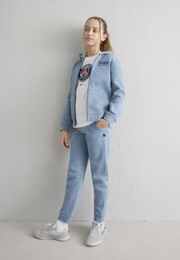 Fată tânără stând lângă un perete alb, purtând un trening albastru deschis, un tricou alb Paris Saint-Germain și adidași gri.