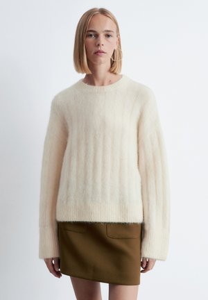 Cremiger, gerippter Pullover aus weichem, strukturiertem Stoff, kombiniert mit einem braunen Rock mit zwei Vordertaschen. Der Pullover hat einen runden Ausschnitt.