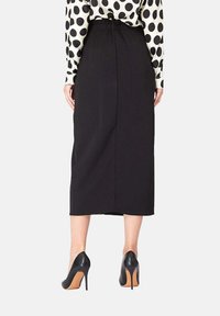 Jupe noire mi-longue avec une taille élastique, vue de dos, associée à des chaussures à talons hauts noires et un haut à pois.