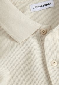 Polo en maille côtelée couleur crème avec patte de boutonnage et col, avec une étiquette "JACK & JONES" à l'intérieur au niveau de la nuque.