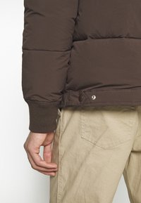 Brązowa kurtka puchowa o pikowanej fakturze, z prążkowanymi mankietami i zatrzaskami u dołu, noszona na jasnobeżowych, teksturowanych spodniach.