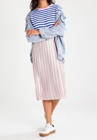 Kvinna som bär en blårandig skjorta, en ljus denimjacka, en rosa plisserad midikjol och vita sneakers, stående med armarna i kors.