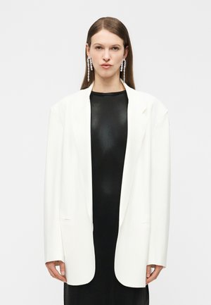 Mujer con blazer blanco largo sobre vestido negro, con pendientes largos de cristal en forma de gota, posando frente a un fondo claro liso.