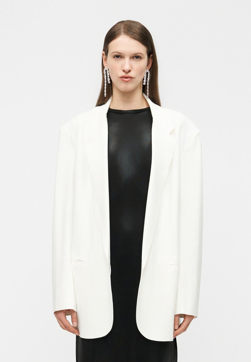 Γυναίκα που φοράει μακρύ λευκό σακάκι πάνω από μαύρο φόρεμα, με μακριά σκουλαρίκια κρύσταλλα σε σχήμα σταγόνας, στέκεται μπροστά σε απλό φωτεινό φόντο.