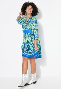 Vestido multicolorido con patrones en azul y verde, mangas tres cuartos, cintura ceñida con un cinturón azul y una falda acampanada, combinado con botas tobilleras plateadas.