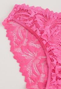 Rozā mežģīņu apakšveļa ar ziedu rakstiem, viļņotām malām un gludu rozā jostas daļu. Mežģīņu dizains ir izsmalcināts ar teksturētu virsmu.