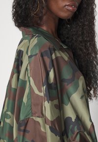 Donna con lunghi capelli ricci che indossa un giubbotto a motivi camouflage verde, marrone e nero, mostrando il profilo laterale del viso e della parte superiore del corpo.