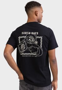 Μαύρο κοντομάνικο μπλουζάκι με λευκό σχέδιο που απεικονίζει τον Άγιο Βασίλη να κρατά μια κυκλική σφραγίδα έγκρισης, με καρό περίγραμμα και κείμενο από πάνω.