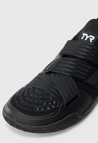 Close-up of black sports shoe with textured knit upper, two wide adjustable straps, and a rubber sole with patterned tread.