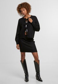Černý pletený cardigan s předním zavazováním, v kombinaci s černou těsnou sukní. Modelka má na sobě černé kožené boty po kolena. Jednoduchý, texturovaný design.