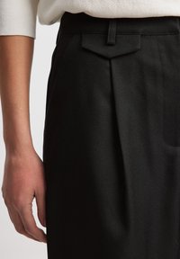 Černé kalhoty na míru s texturovanou látkou, vybavené bočními sklady, úzkým střihem a V-řízným zadním sedlem. Ruka spočívající na látce.
