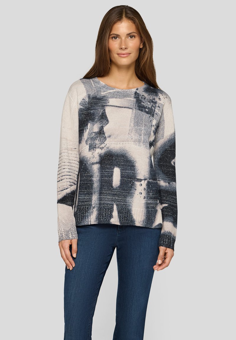 Gestrickter Pullover in Hellbeige und Dunkelblau mit abstrakten Mustern, mit Rundhalsausschnitt und langen Ärmeln, kombiniert mit dunkelblauer Jeans.