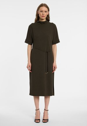 Vrouw draagt een donker olijfgroene midi-jurk met korte mouwen, een hoge halslijn en een dunne riem gestrikt in de taille, gecombineerd met zwarte open-toe hakken.