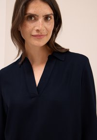 Frau mit schulterlangem braunem Haar und Sommersprossen, die eine dunkelmarineblaue V-Ausschnitt-Bluse vor einem schlichten hellen Hintergrund trägt.