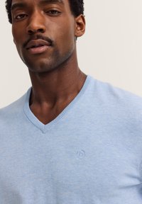 Nahaufnahme eines Mannes in einem hellblauen V-Ausschnitt-Pullover mit einem kleinen gestickten Logo auf der Brust vor einem schlichten Hintergrund.