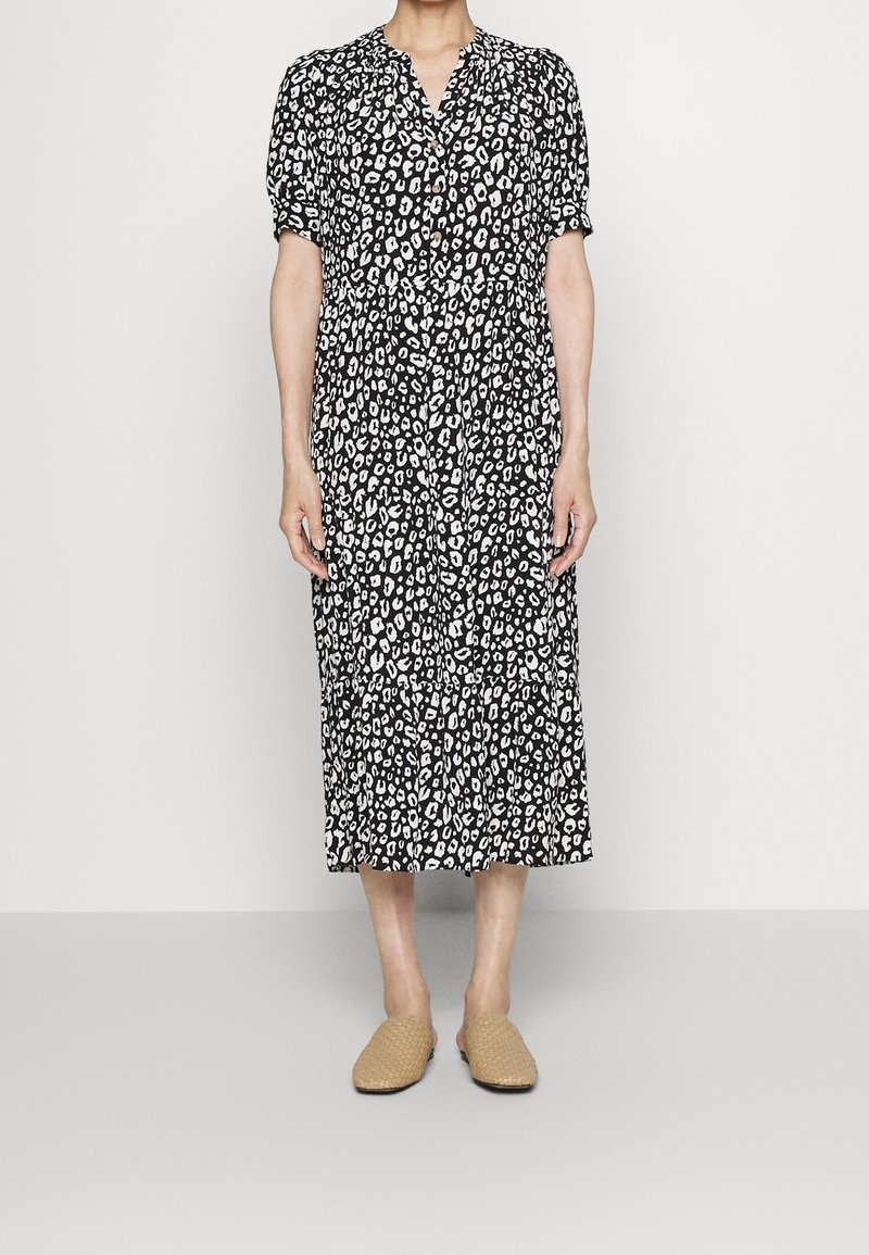 Schwarz-weißes Leopardenmusterkleid mit kurzen Ärmeln, einem V-Ausschnitt und einem schichtweisen Rock. Getragen mit hellbraunen, geflochtenen Slippers.