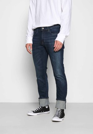 Ein Mann trägt hochgekrempelte dunkelblaue Jeans, schwarze High-Top-Sneaker und ein langärmeliges weißes Hemd und steht vor einem einfachen Hintergrund.