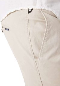 Beige katoen broek met een zijzak met een zwarte rits, gestikte details en een klein merkje op de zak.