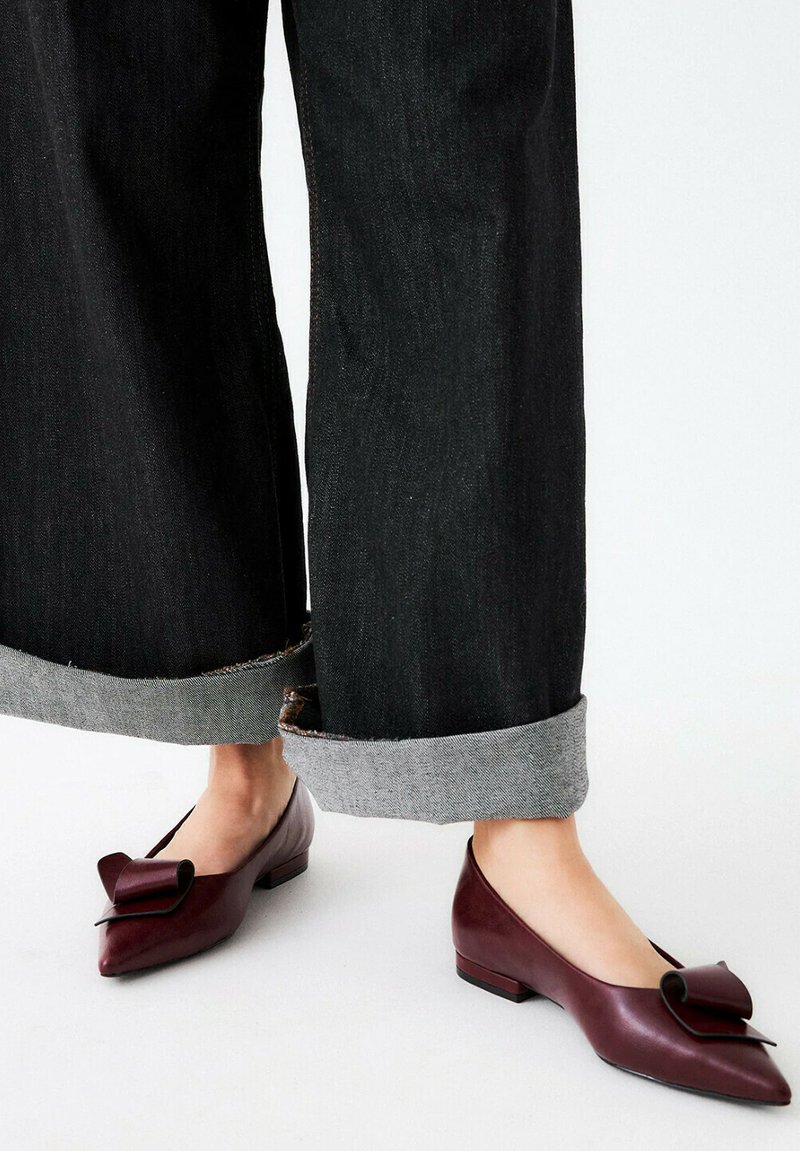 Scarpe basse in pelle color bordeaux con punta affilata e fiocco decorativo. Indossate con jeans neri a gamba larga con orli grezzi e risvolti grigi.